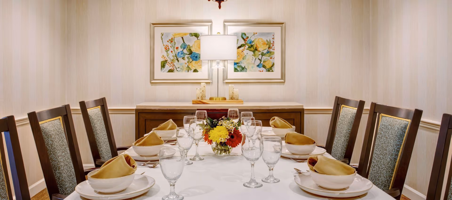 A formal dining room table set for eight with white tablecloth, beige napkins folded in bowls, multiple glasses, and silverware. A small floral centerpiece with yellow, red, and white flowers is in the middle. Behind the table is a wooden sideboard with a glass lamp and two framed floral paintings on the wall.