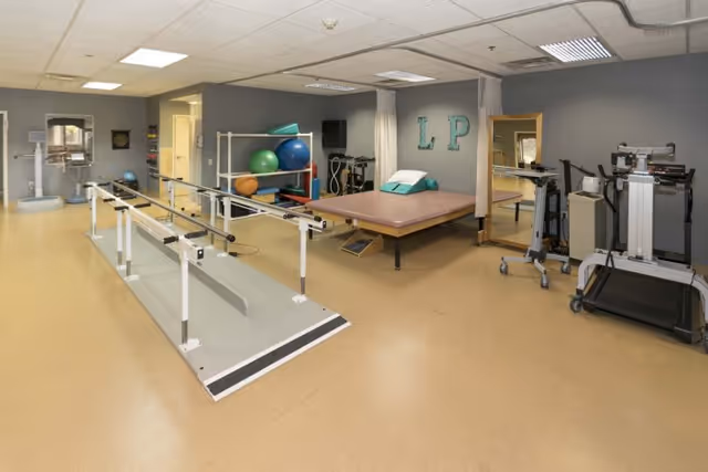 A rehabilitation therapy room with parallel bars for walking exercises, a large padded therapy table, exercise balls on a shelf, a treadmill, and other physical therapy equipment. The walls are gray with the letters 'L P' mounted on one wall, and there is a large mirror beside the therapy table.