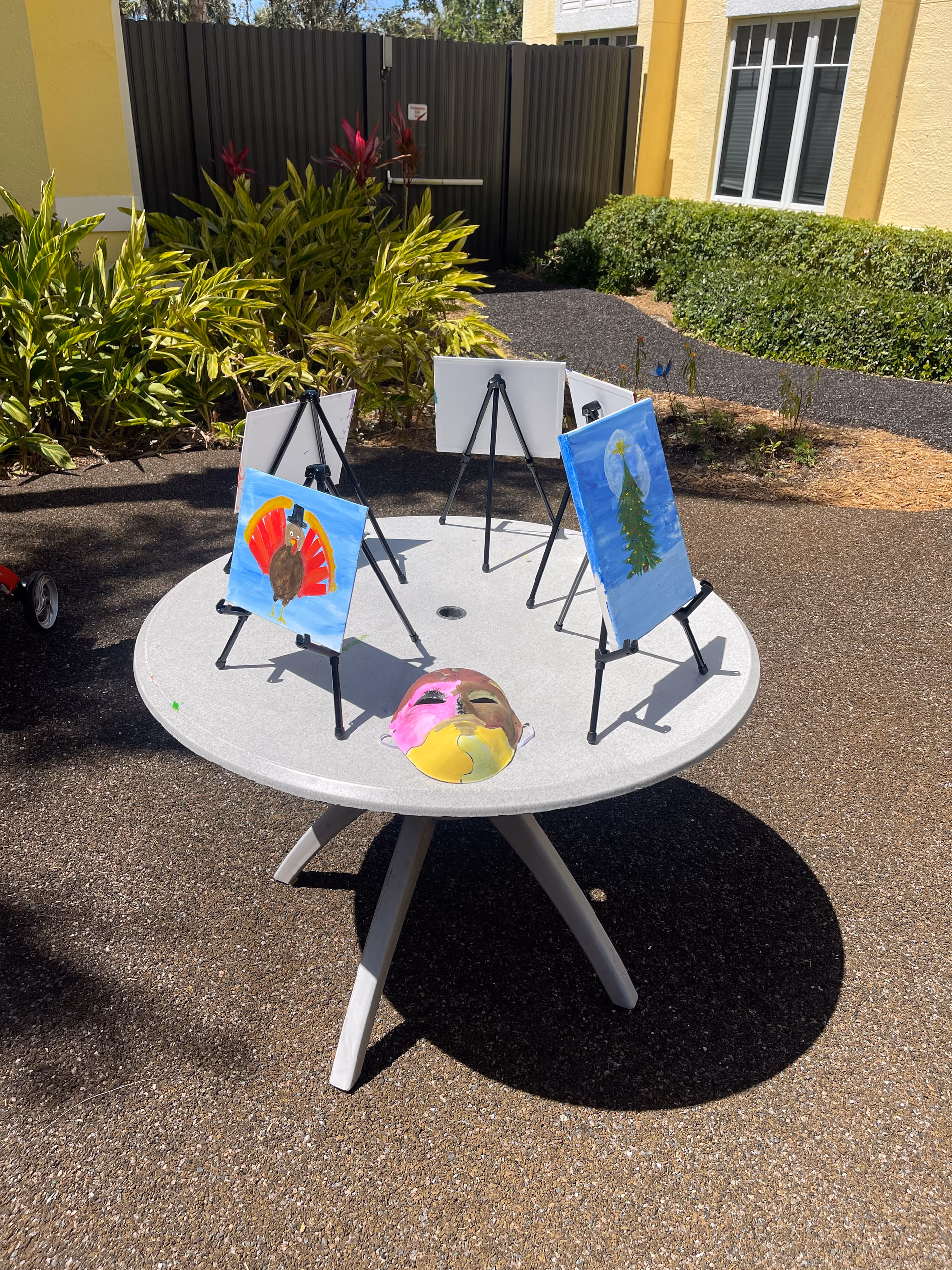 A round outdoor table with four small easels holding paintings, including a turkey and a Christmas tree, and a colorful mask placed on the table. The table is set on a paved area with plants and a building with windows in the background.