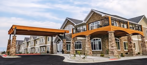 Front exterior of a two-story senior living building with a covered porte-cochere supported by stone columns.