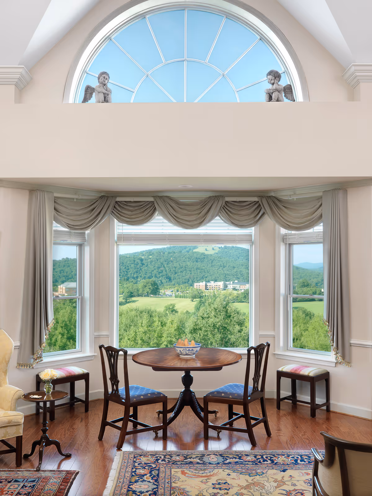 A cozy sitting area with a round wooden table and two wooden chairs with blue cushions, positioned in front of large windows that offer a scenic view of green hills and trees. The windows are adorned with beige curtains and a decorative valance. Above the windows is a large arched window with two small angel statues on the ledge. The room has hardwood floors and a patterned area rug, with additional seating including a cushioned bench and an armchair.