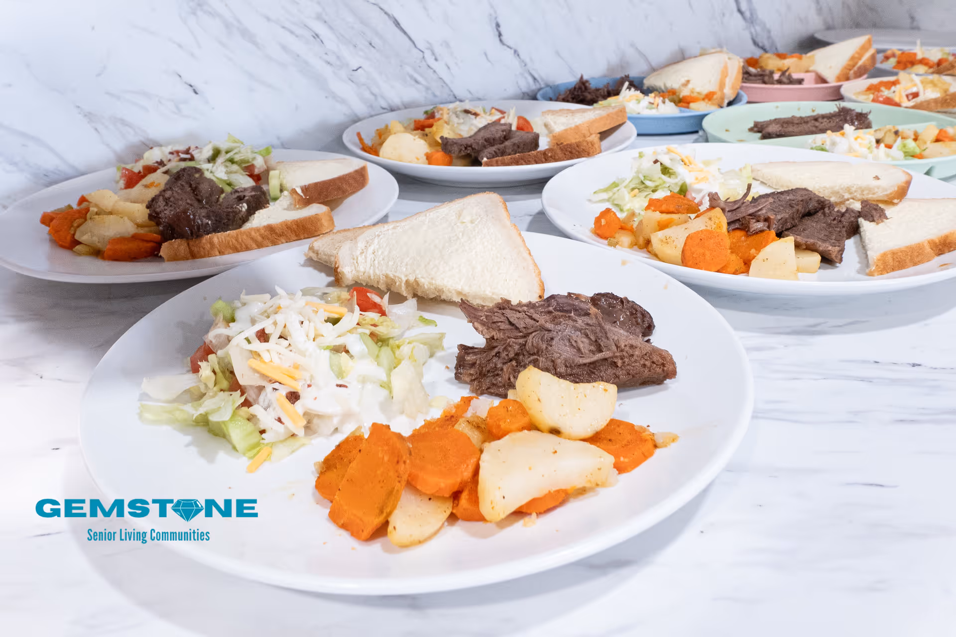 Several white plates arranged on a marble countertop, each containing a meal with cooked carrots and potatoes, a portion of shredded beef, a slice of white bread, and a side salad topped with shredded cheese and dressing.