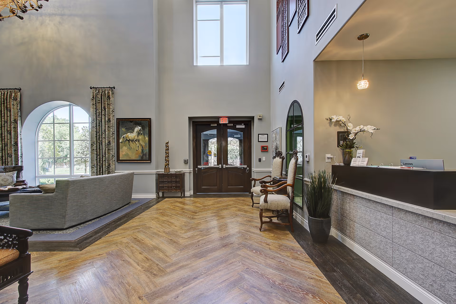 Interior view of a senior living facility lobby with a wooden double door exit, high ceiling, large windows with curtains, a seating area with a gray sofa, two upholstered armchairs, a reception desk with a computer and a vase of white flowers, and decorative wall art.