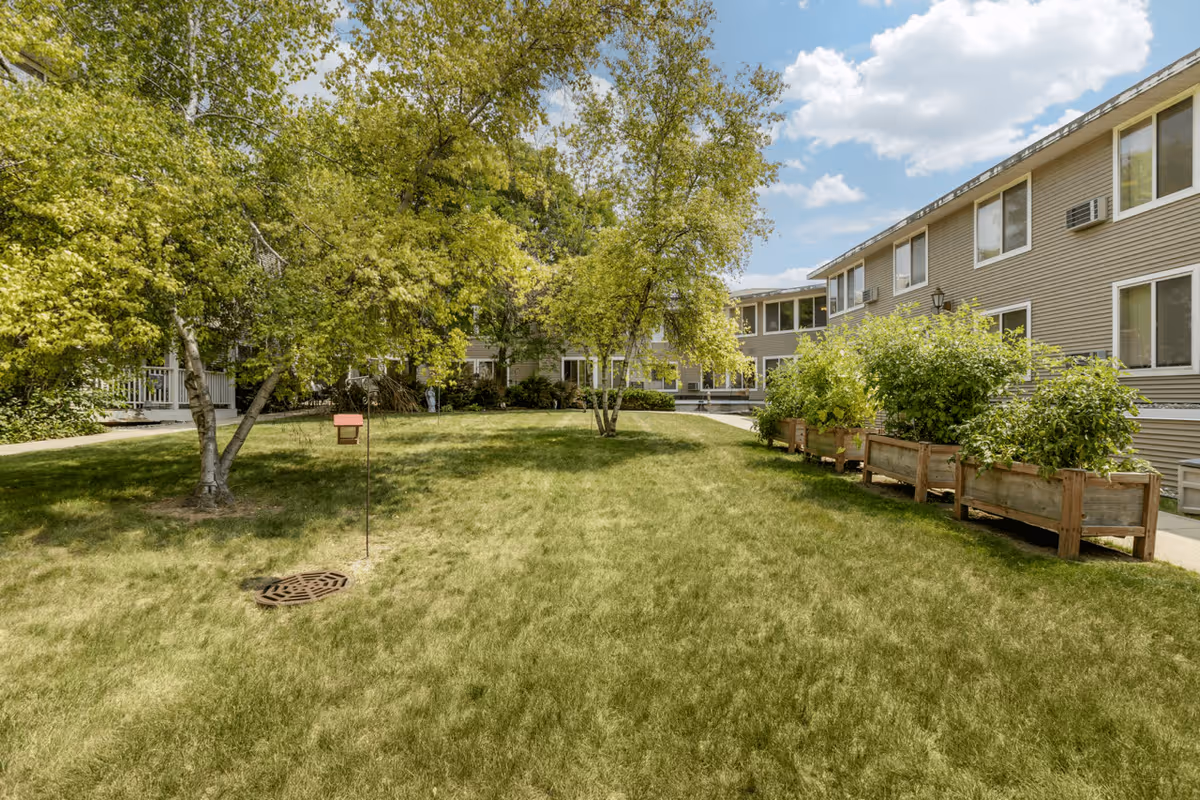 A grassy courtyard area at Layton Terrace Senior Living with several trees and raised garden beds along a paved walkway. The courtyard is surrounded by a two-story building with multiple windows and air conditioning units. The sky is partly cloudy.