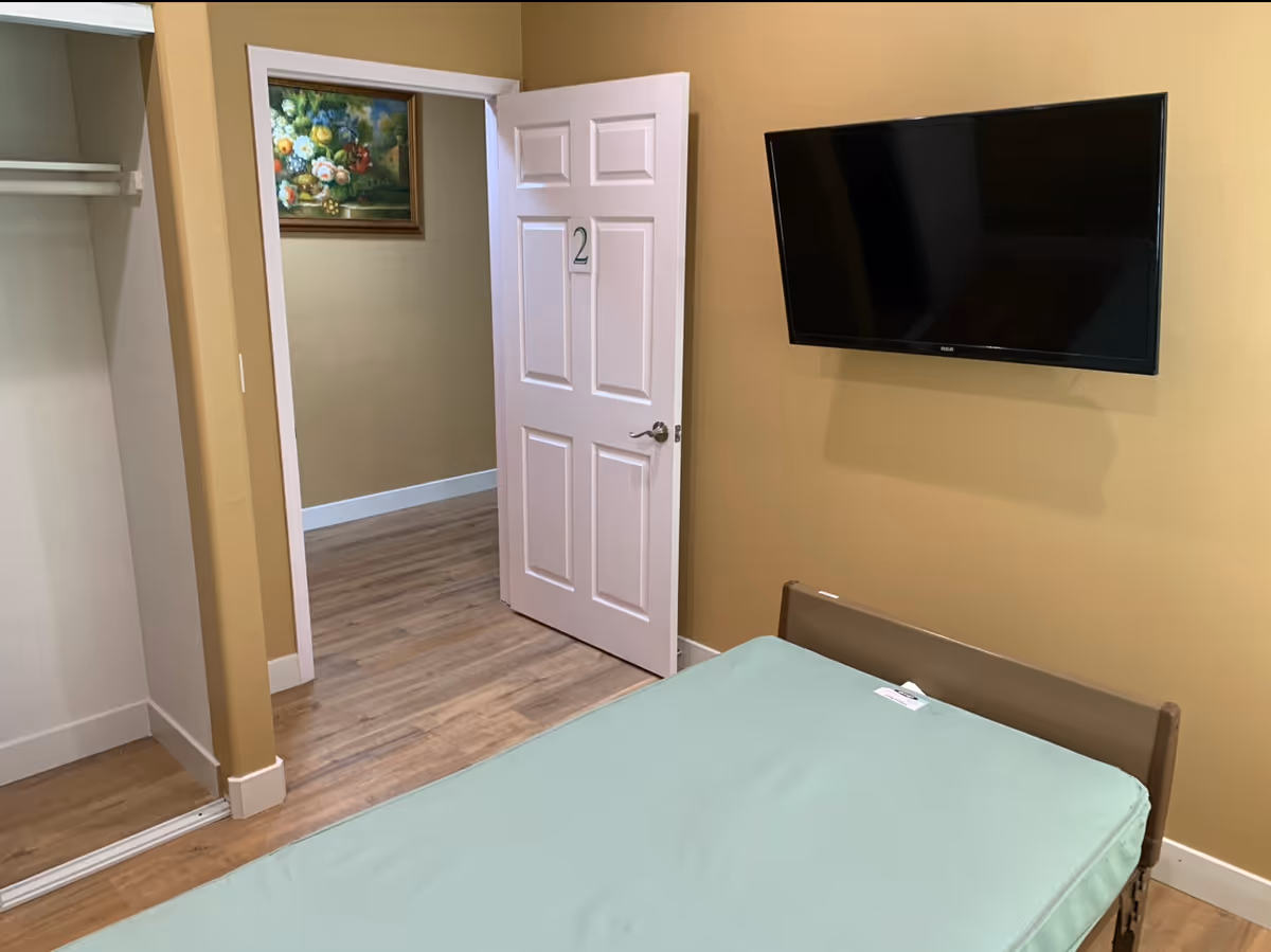 A simple bedroom with a single bed covered with a light green sheet, a wall-mounted flat-screen TV, an open white door labeled with the number 2, and a painting of flowers on the wall in the hallway outside the room. The walls are painted beige and the floor has wood-like vinyl flooring.