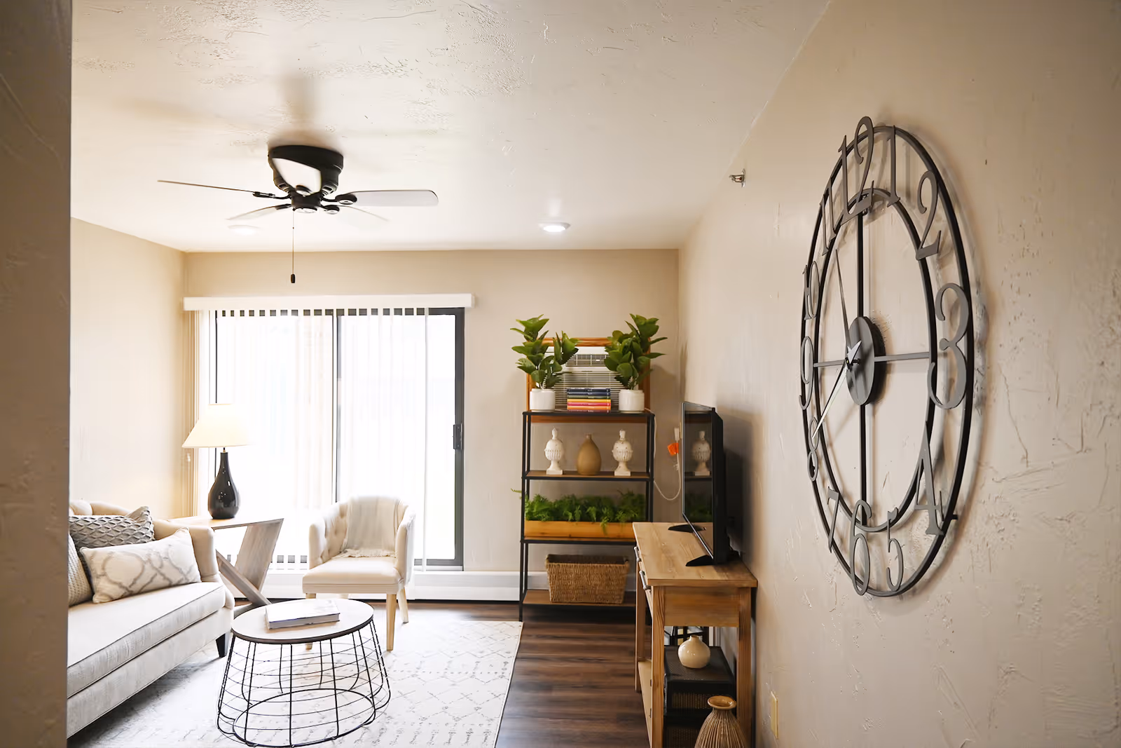 Bright furnished living room with a sofa, armchair, round coffee table, shelving unit, TV stand, and a large wall clock by sliding glass doors.