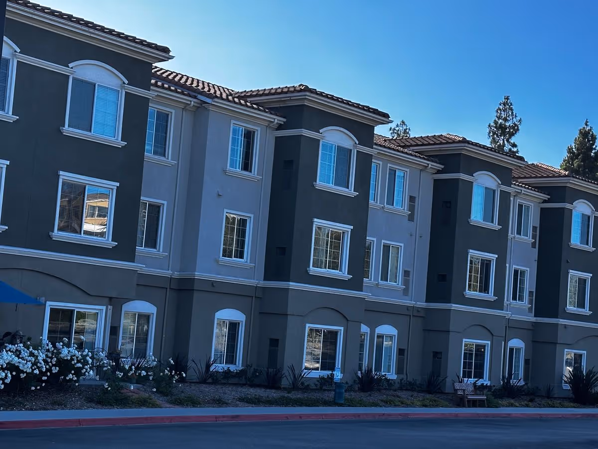 Front exterior of a three-story stucco building with tiled roof, multiple windows and landscaped beds.
