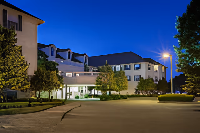 Front exterior of a multi-story senior living building at dusk with an illuminated entrance, driveway, and landscaped trees.