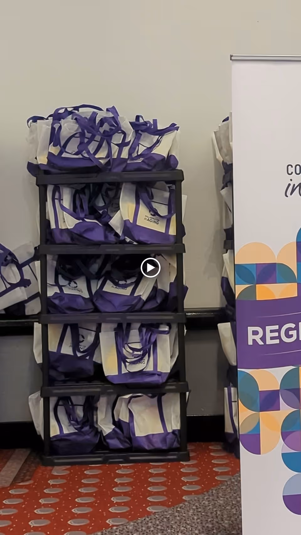 A black shelving unit filled with multiple white and purple tote bags stacked on each shelf. To the right, there is a partial view of a colorful banner with geometric shapes and the word 'REGISTER' visible. The floor has a red carpet with circular patterns.