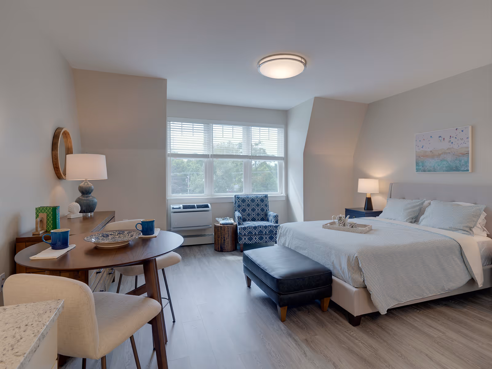 A bright and cozy bedroom in a senior living facility featuring a large bed with white and light blue bedding, a dark blue ottoman at the foot of the bed, a blue patterned armchair near a window with white blinds, a wooden table with two blue mugs and chairs, and two lamps providing warm lighting.