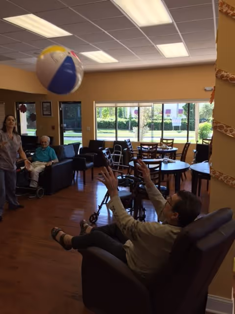 People in a senior living common room sitting in chairs and tossing a large beach ball near tables and windows.