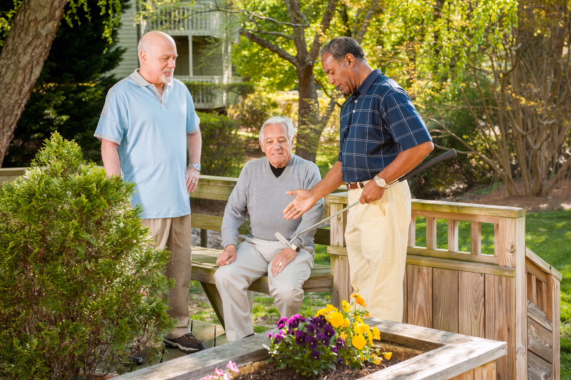 Three elderly men outdoors in a garden area at a retirement community. One man is sitting on a wooden bench while the other two stand nearby. One standing man is holding a golf club and gesturing as if explaining something. There are trees, bushes, and flowers around them, and a wooden railing behind the bench.
