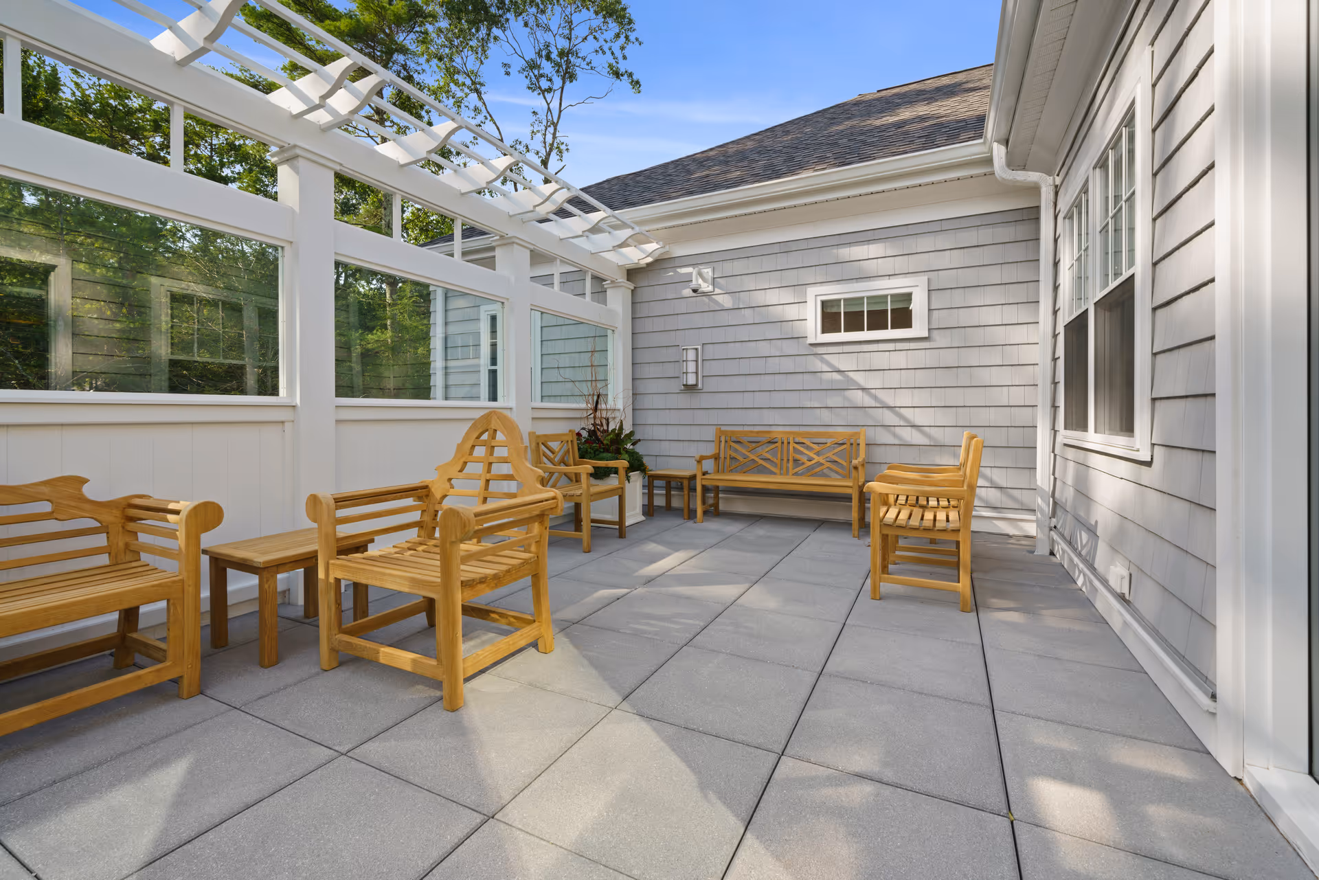 Outdoor patio area with wooden benches and chairs arranged on a tiled floor, surrounded by white fencing and a pergola overhead, attached to a gray building with windows.