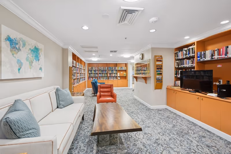 A cozy library or reading room with built-in orange bookshelves filled with books, a beige sofa with blue cushions, a wooden coffee table, and two armchairs, one red and one blue. The room has a patterned carpet, a world map artwork on the wall, and a TV on one of the shelves.