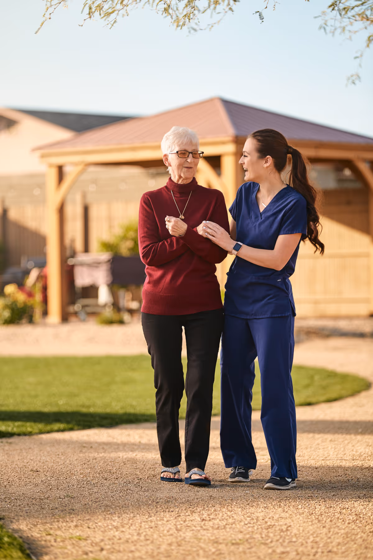 An elderly woman wearing a red sweater and black pants is walking outdoors on a gravel path, supported by a smiling caregiver dressed in blue scrubs. They are in a garden area with a wooden gazebo and greenery in the background.