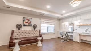 A spacious, well-lit room with a tufted bench against the wall, two small white stools, a large window with blinds, and a built-in counter with a mirror and a swivel chair. The room has wooden flooring, recessed ceiling lights, and neutral-colored walls with minimal decor including a framed picture and two decorative fans on the bench.