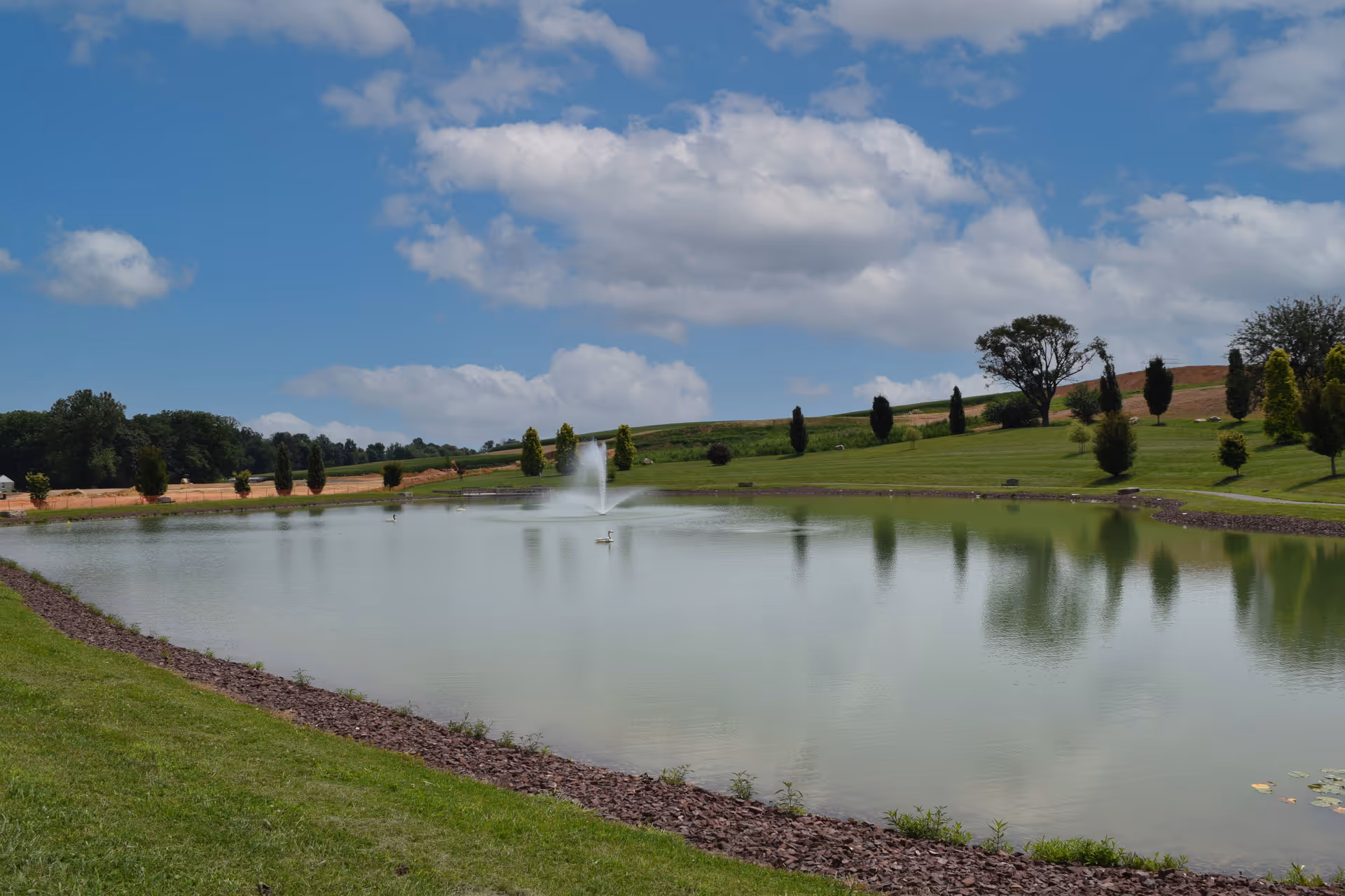 A man-made pond with a central fountain surrounded by grassy lawns, trees, and a partly cloudy blue sky.