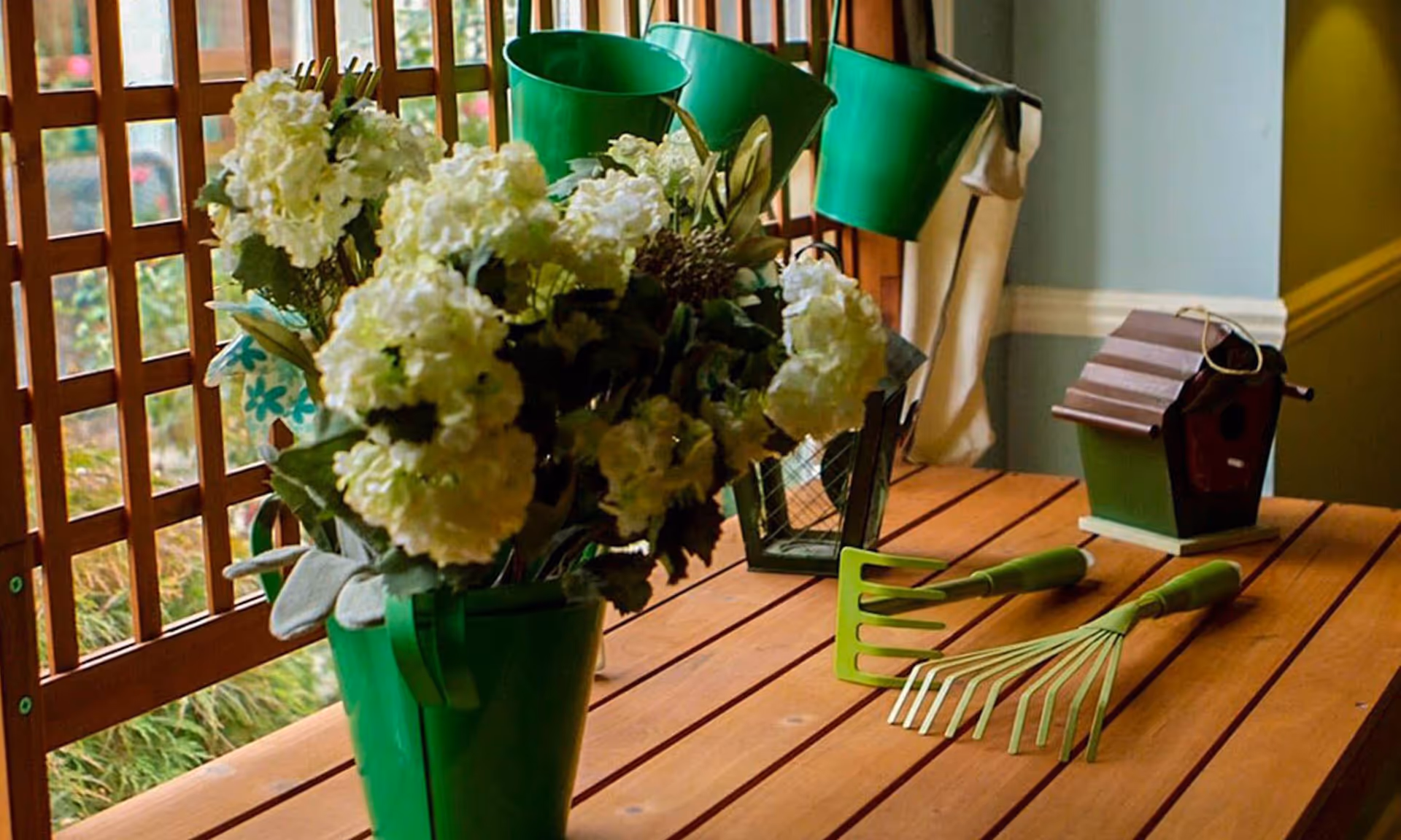 A wooden table with green gardening tools including a small rake and a hand cultivator, a green bucket holding white flowers, a small birdhouse, and a lantern. The background shows a wooden lattice and part of an indoor wall.