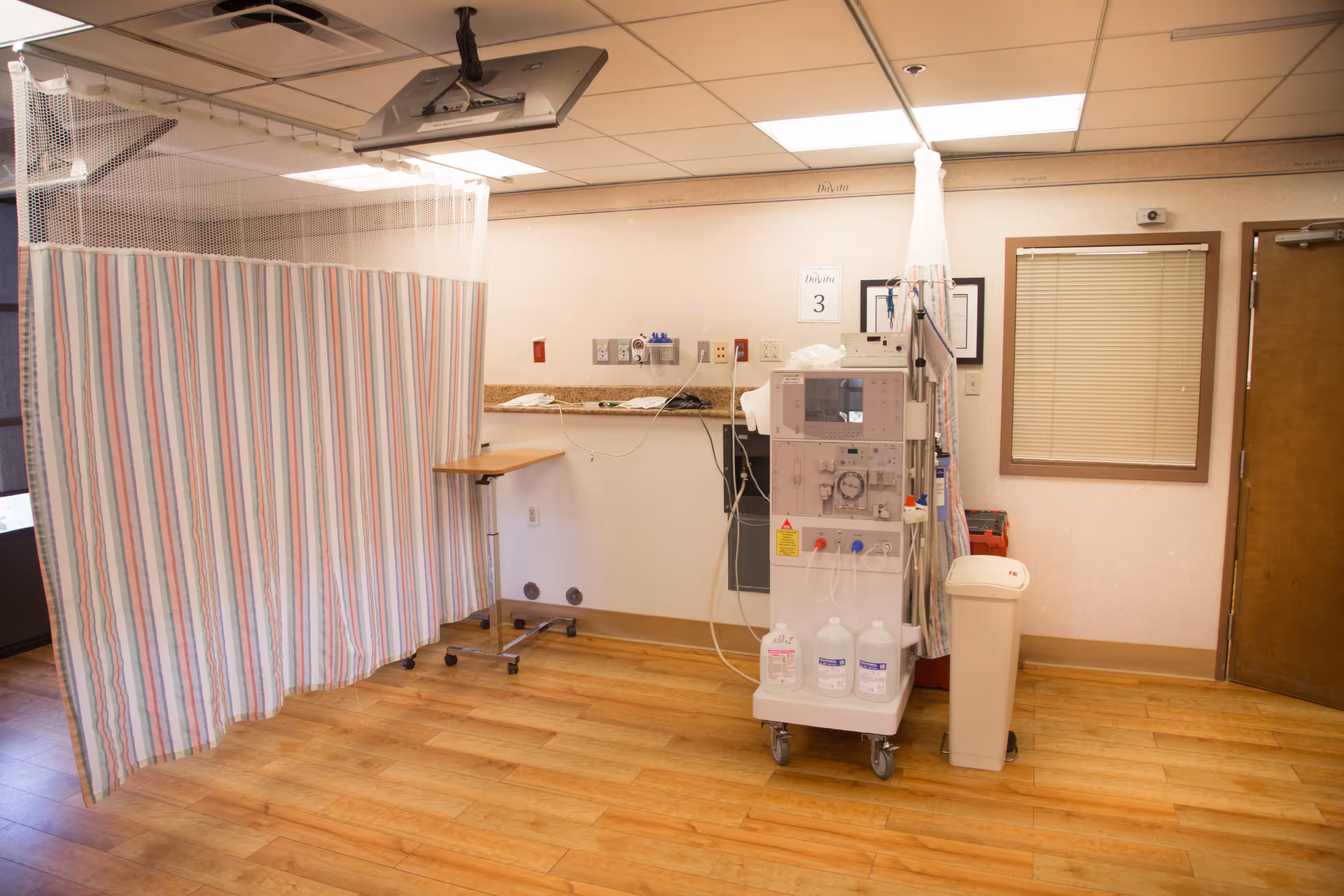 A medical treatment room with a dialysis machine, a privacy curtain with vertical stripes, a small adjustable table on wheels, a trash bin, and a window with closed blinds. The room has wooden flooring and a drop ceiling with fluorescent lighting.