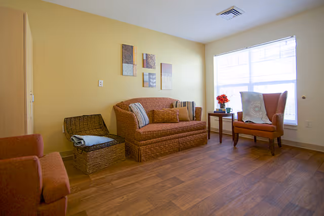 A cozy living room with a patterned sofa, an armchair with a throw blanket, a wicker basket with a folded blanket, a small side table with a vase of red flowers, and a large window letting in natural light. The walls are painted light yellow and the floor is wooden.
