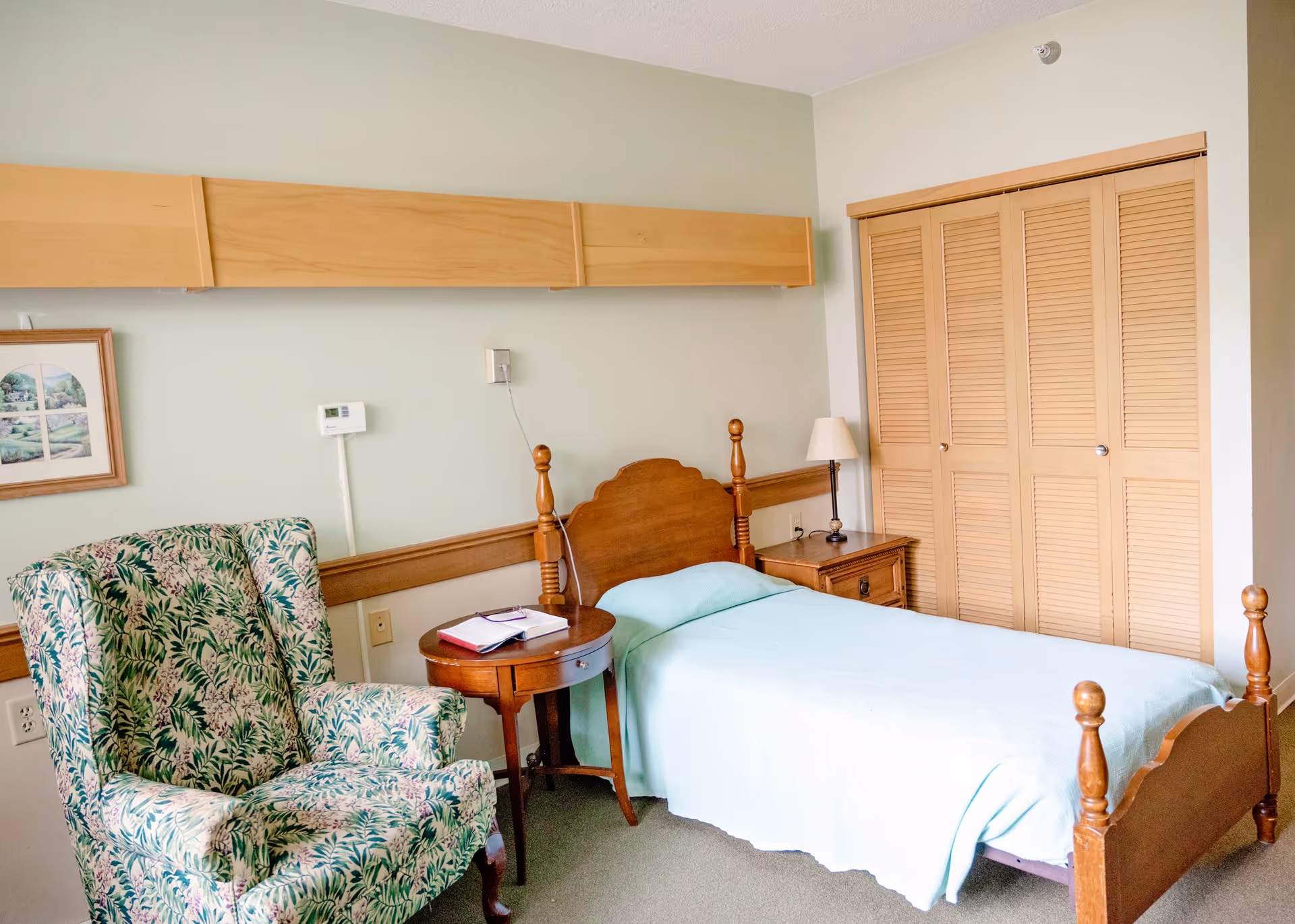 A small bedroom with a single wooden bed covered with a light blue blanket. Next to the bed is a wooden nightstand with a lamp and a round wooden side table with a book and glasses on it. There is a floral patterned armchair to the left and a closet with wooden louvered doors on the right. The walls are light green with a framed picture hanging on the left side.