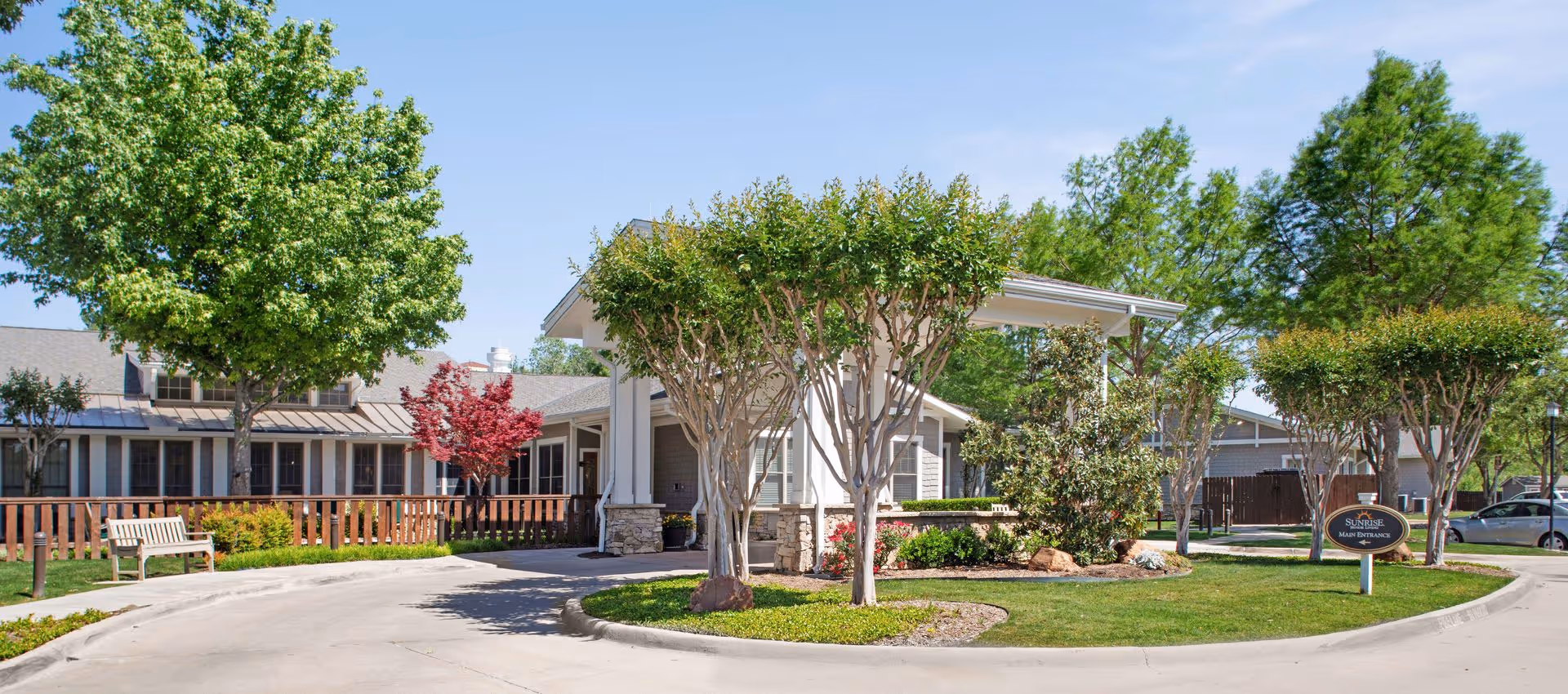 Exterior view of Sunrise of Fort Worth senior living facility entrance with a circular driveway, manicured landscaping including trees and bushes, a wooden bench, and a sign indicating the main entrance on a sunny day.