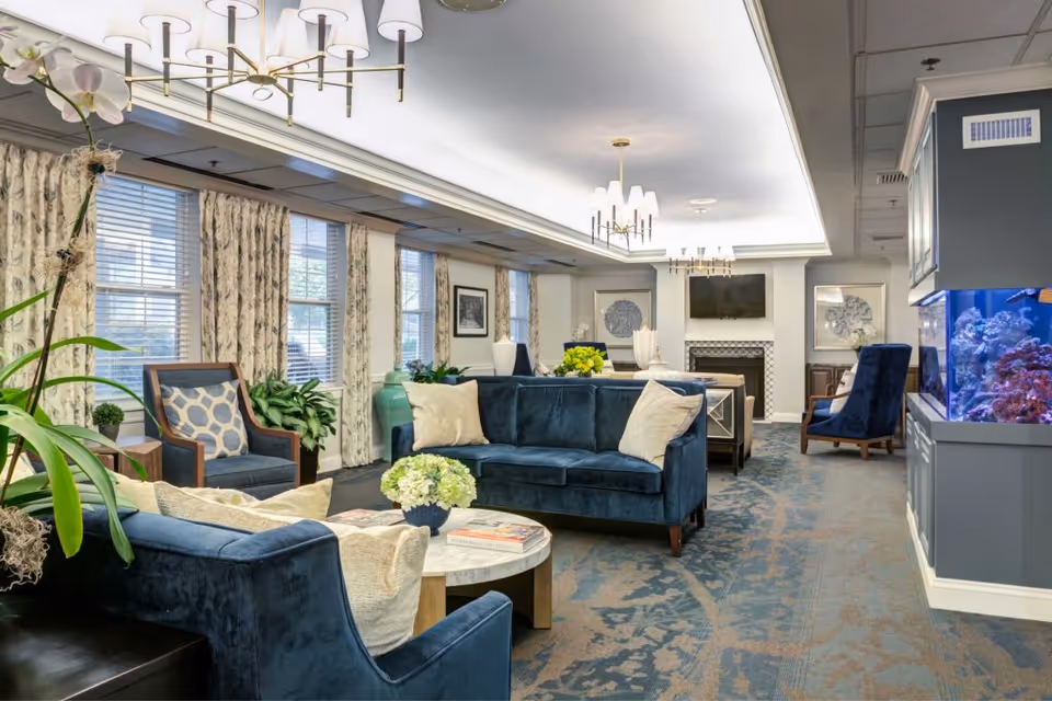 A well-lit living room with blue velvet sofas and armchairs, a round coffee table with a flower arrangement and magazines, large windows with patterned curtains, a fireplace with a mounted TV above it, framed artwork on the walls, and a large built-in aquarium on the right side.