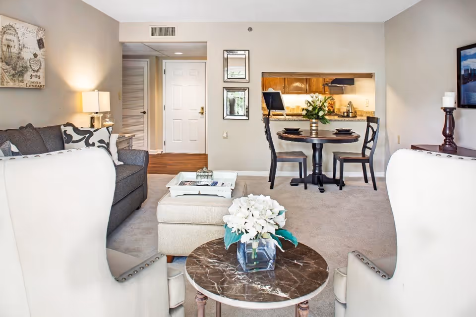 A cozy living and dining area in a senior living facility. The room features two white armchairs facing a small round marble-top table with a vase of white flowers. Behind the table is a beige ottoman with a tray on top, and a gray sofa with patterned pillows. A small round dining table with two chairs is set near a pass-through window to the kitchen. The walls are light-colored with framed artwork and mirrors, and a lamp provides warm lighting.