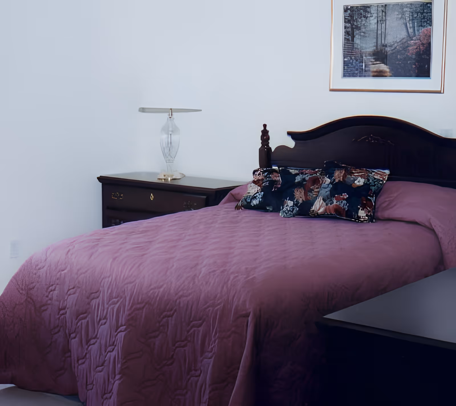 A bedroom with a dark wooden bed frame and a purple bedspread. There are two decorative pillows with a floral pattern on the bed. Next to the bed is a dark wooden nightstand with a clear glass lamp. A framed picture of a forest path hangs on the white wall above the bed.