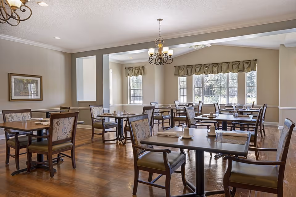A bright dining room with several wooden tables and chairs arranged neatly. Each table has place settings including cups, glasses, and napkins. Large windows with valance curtains allow natural light to fill the room. The floor is wooden, and there are two chandeliers hanging from the ceiling. A framed picture is on the wall.