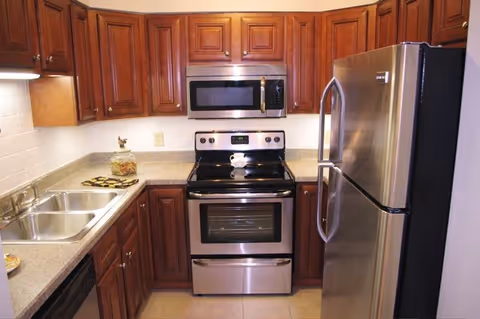 A kitchen with wooden cabinets, a stainless steel refrigerator, an electric stove with oven, a microwave above the stove, a double sink, and a countertop with a decorative jar and a small plate.