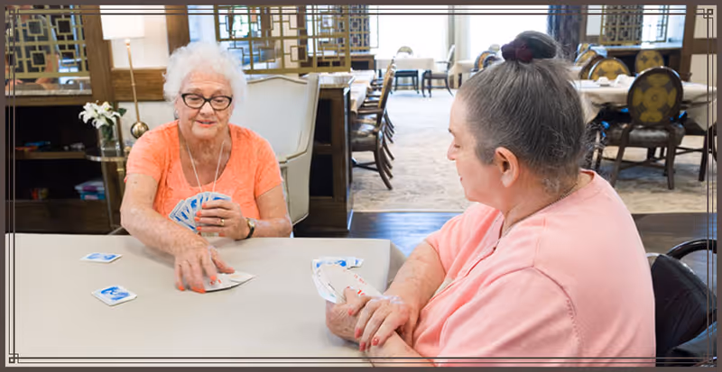 Two elderly women sitting at a table playing cards in a well-lit room with chairs and tables in the background.