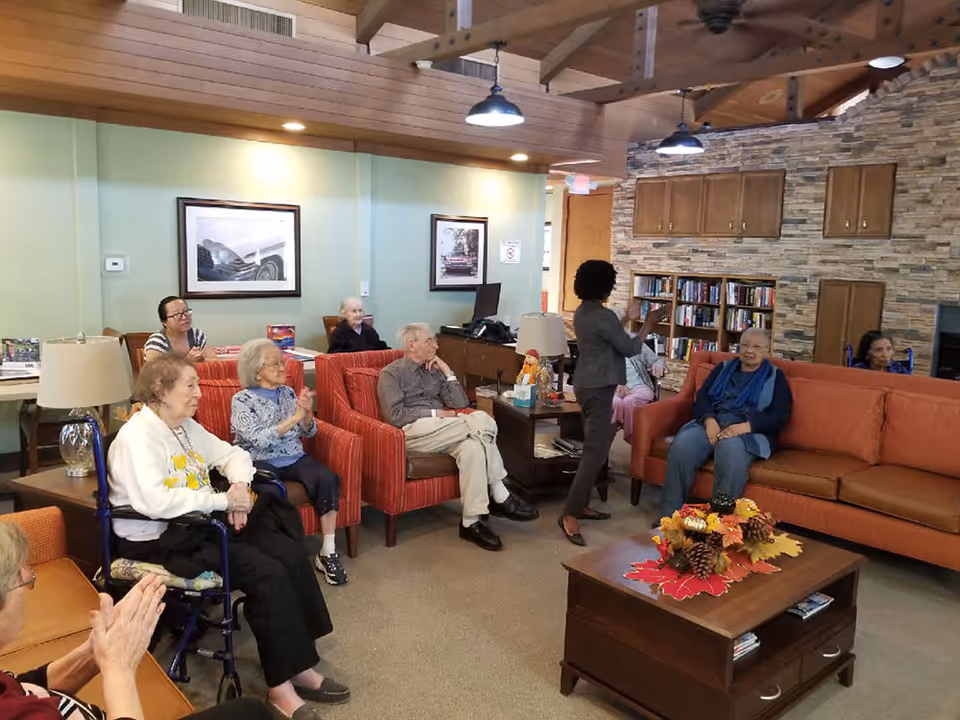 A group of elderly people seated in a cozy living room area with a woman standing and speaking to them. The room features comfortable chairs and sofas, a wooden coffee table with autumn-themed decorations, bookshelves, framed pictures on the wall, and a stone accent wall with wooden cabinets.