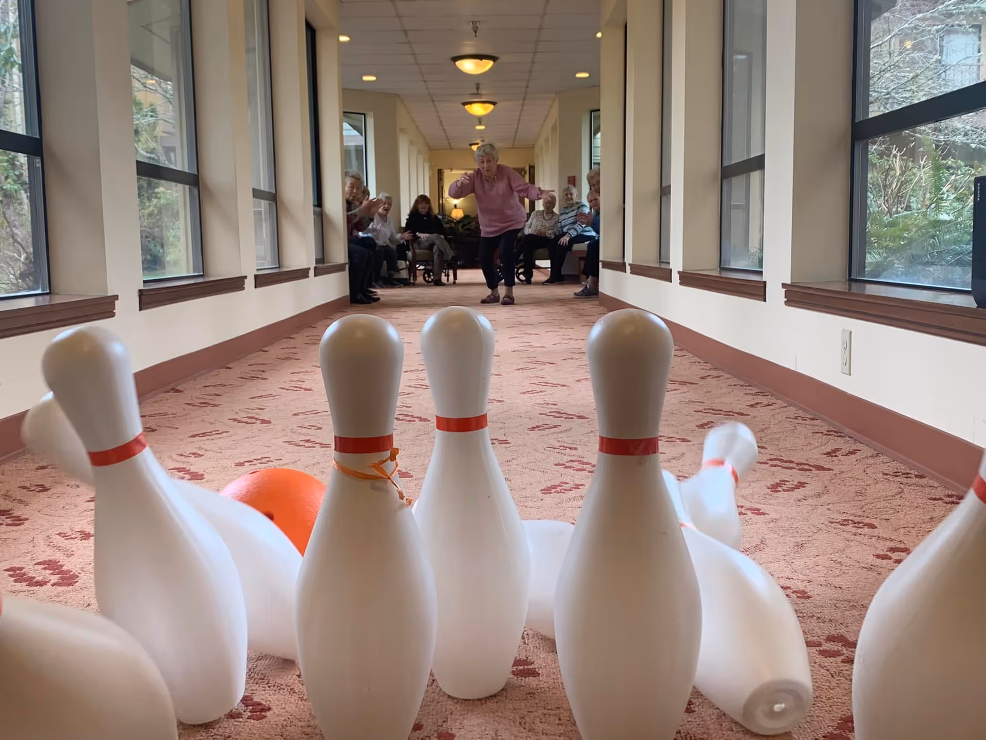 An elderly woman is bowling with a bright orange ball in a carpeted hallway lined with large windows. Several other elderly people are seated along the sides of the hallway, watching and cheering her on.