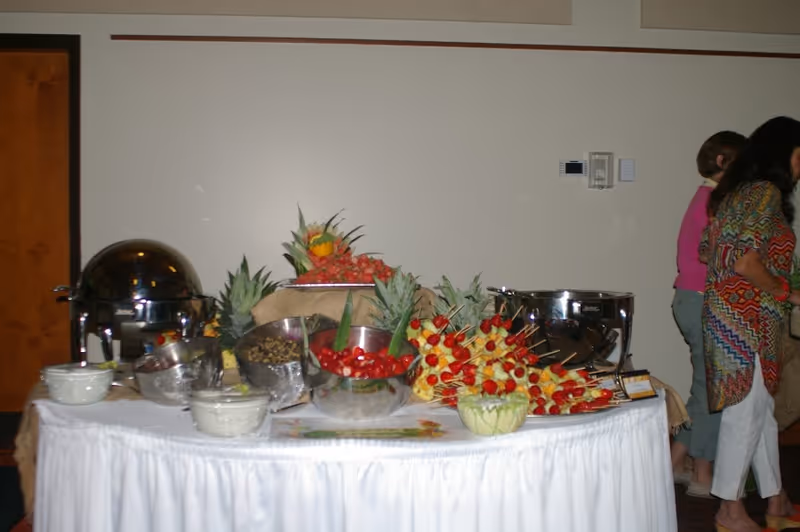 A buffet table set up with various bowls of food including fruit skewers, a bowl of cherry tomatoes, and other dishes. The table is covered with a white tablecloth and decorated with pineapples and other fruits. Two women are standing near the table on the right side of the image.