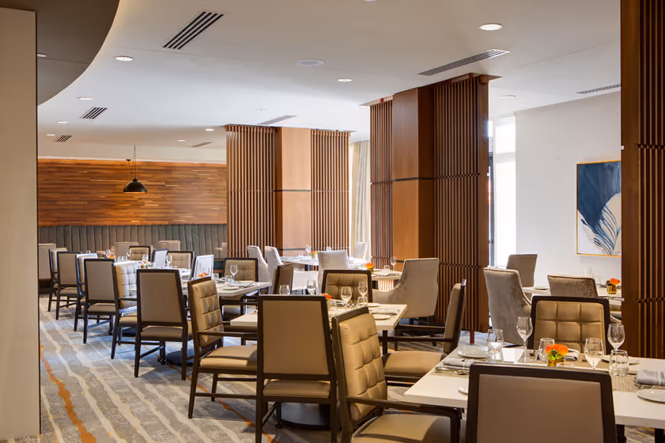 A modern dining room with multiple tables set with plates, glasses, and napkins. The room features cushioned chairs, wooden panel columns, a wooden accent wall, and a carpeted floor with a striped pattern. There is a piece of abstract artwork on the wall and pendant lighting hanging from the ceiling.
