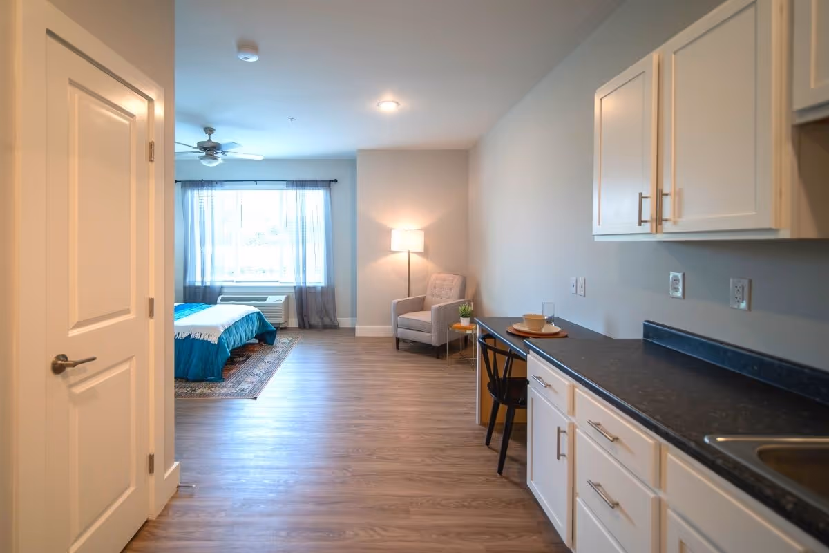 A bright and spacious assisted living apartment featuring a kitchenette with white cabinets and black countertops on the right, a small dining area with a chair and table set with dishes, and a cozy sitting area with an armchair and floor lamp near a large window with sheer curtains. A bed with a blue blanket and a ceiling fan are visible in the background.