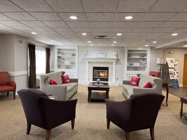 A cozy living room area with two gray sofas facing each other, each with red decorative pillows. Two dark armchairs are positioned in the foreground facing the sofas. A wooden coffee table sits in the center on a beige carpet. In the background, there is a fireplace with a white mantel flanked by built-in shelves holding decorative items. The room has a drop ceiling with recessed lighting and windows with dark curtains on the left side.
