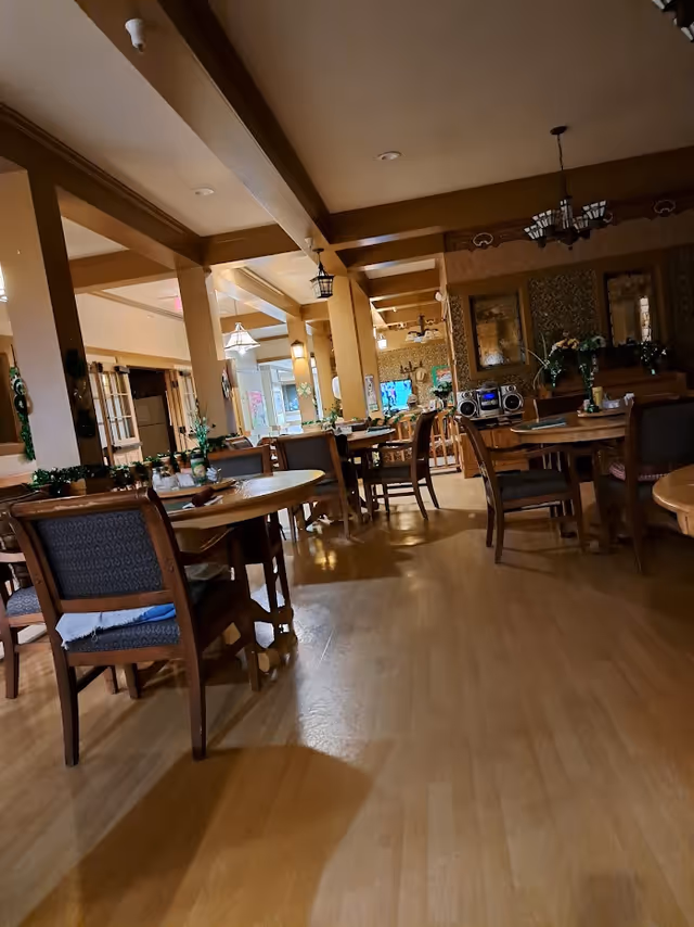 Interior view of a dining room with multiple wooden tables and chairs arranged neatly. The room has wooden flooring, beige walls, and ceiling beams. There are decorative plants on the tables and wall decorations including mirrors and framed art. A stereo system is visible on a side table, and the room is softly lit with ceiling lights and hanging lamps.