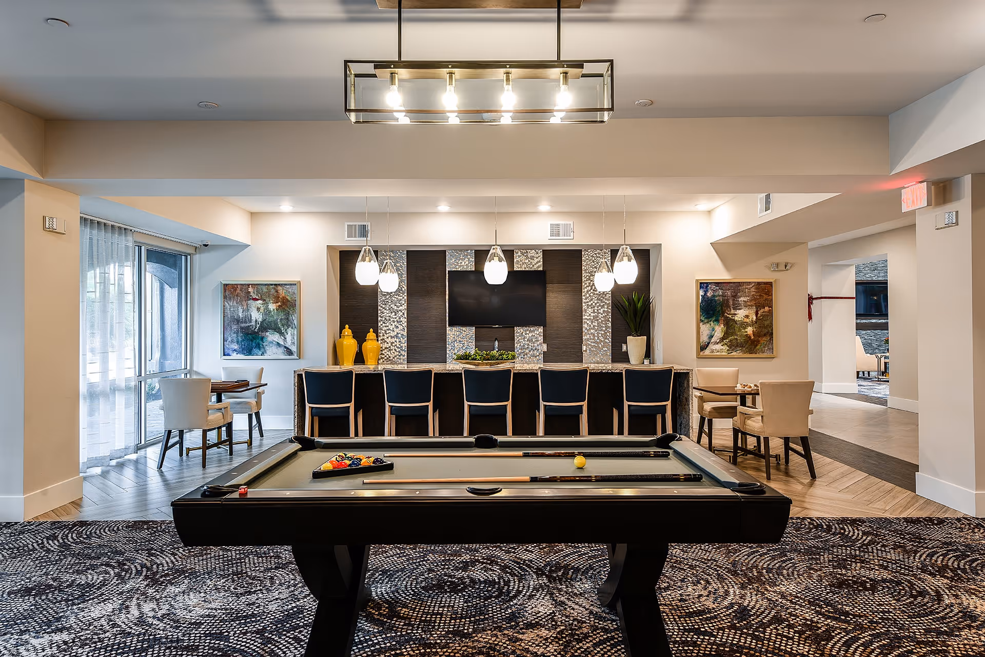 A modern recreational room featuring a pool table in the foreground with pool cues and balls arranged on it. Behind the pool table is a bar area with six high chairs, pendant lights hanging above, decorative yellow vases, a potted plant, and a flat-screen TV mounted on a textured wall. On either side of the room are small seating areas with tables and chairs, and large windows letting in natural light.