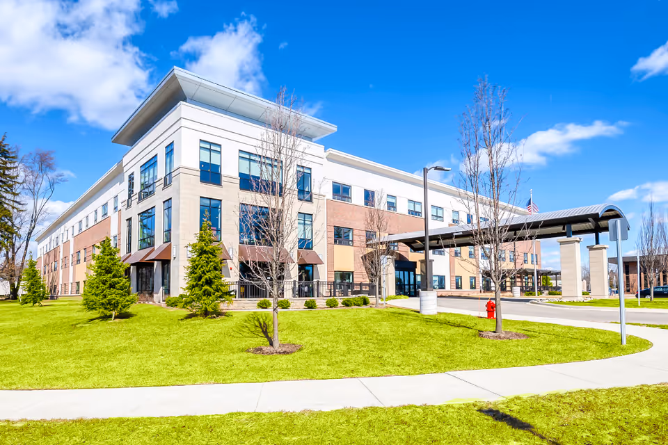 Three-story modern senior living building with a covered entrance, manicured lawn and young trees under a blue sky.