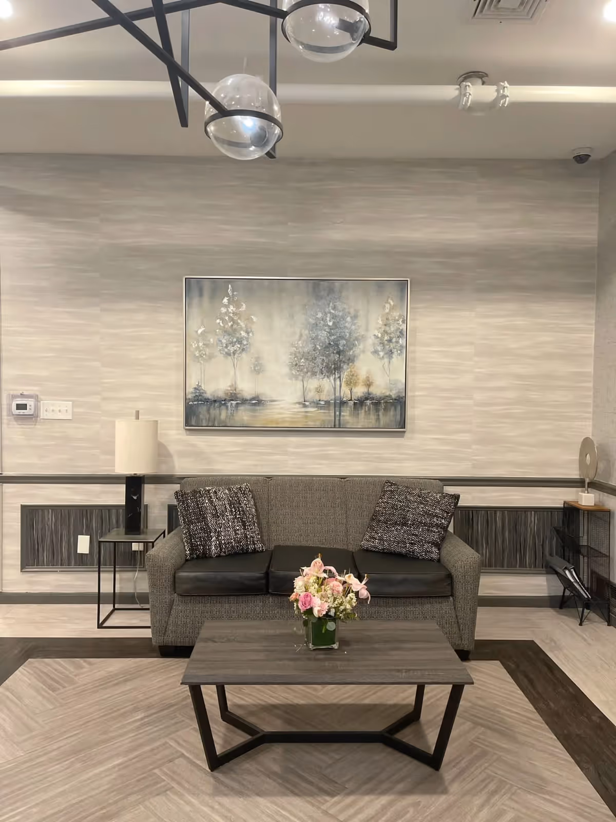 A modern living room area with a gray sofa featuring two patterned cushions, a wooden coffee table with a small flower arrangement in a glass vase, a side table with a lamp, and a large framed painting of trees on the wall behind the sofa. The room has neutral tones with a patterned rug and contemporary lighting fixtures on the ceiling.