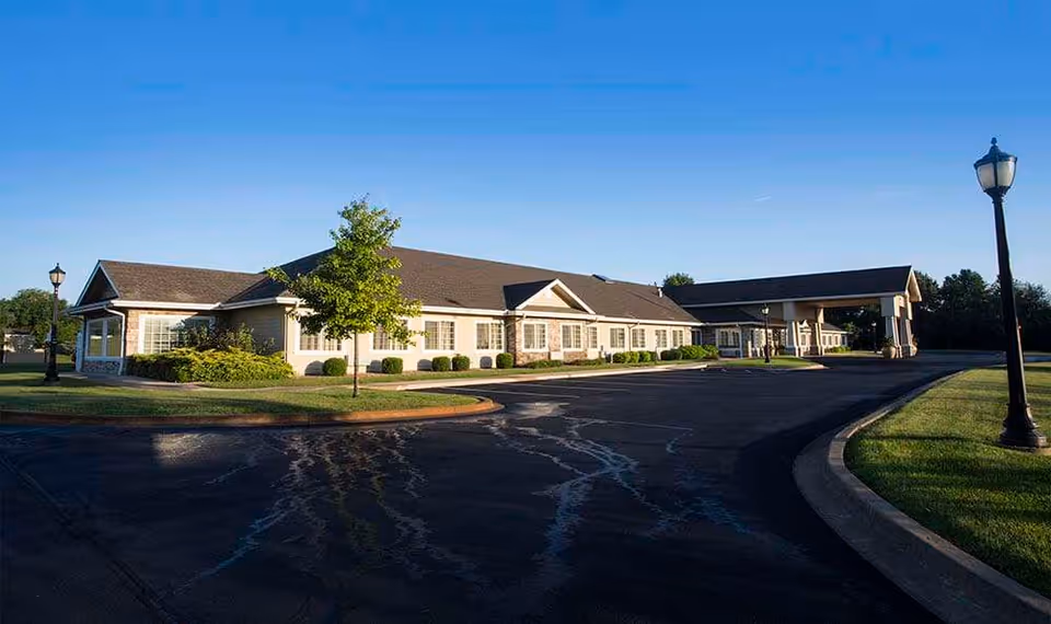 Exterior view of a single-story senior living facility building with a covered entrance, surrounded by a paved driveway, green grass, and a few trees under a clear blue sky.