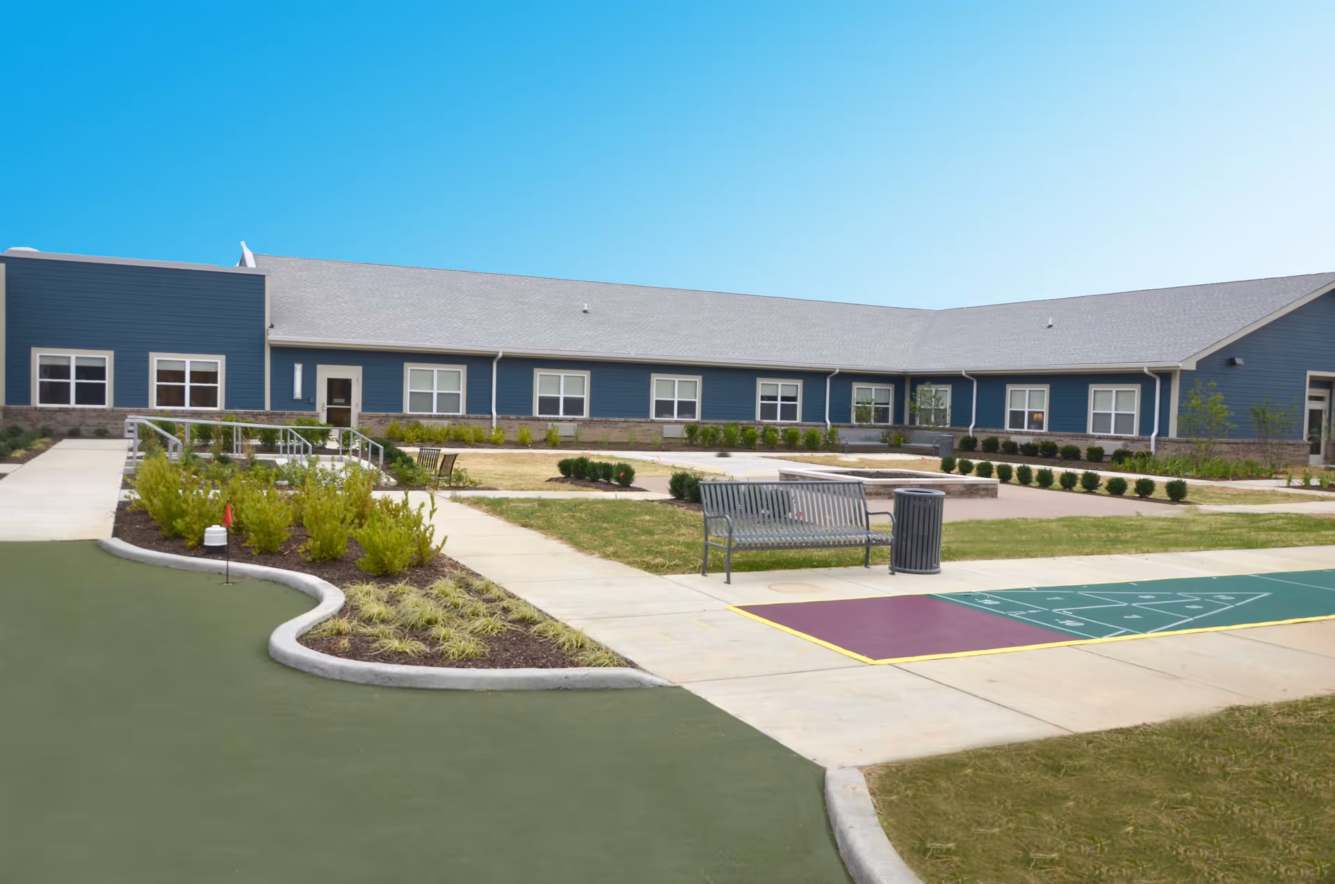 Outdoor courtyard area of Arlington Place Health Campus featuring a shuffleboard court, benches, trash can, landscaped garden beds, and a single-story building with blue siding and multiple windows under a clear blue sky.
