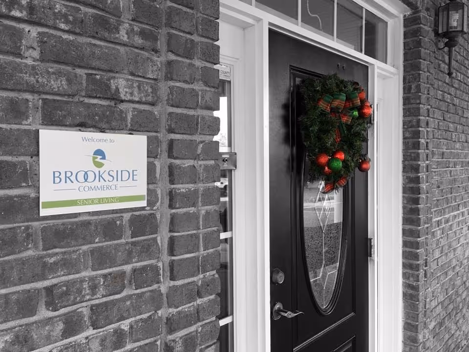 Entrance door of Brookside Commerce senior living facility with a festive wreath decorated with red and green ornaments and a plaid bow hanging on the black door. A white sign on the brick wall next to the door reads 'Welcome to Brookside Commerce Senior Living.'