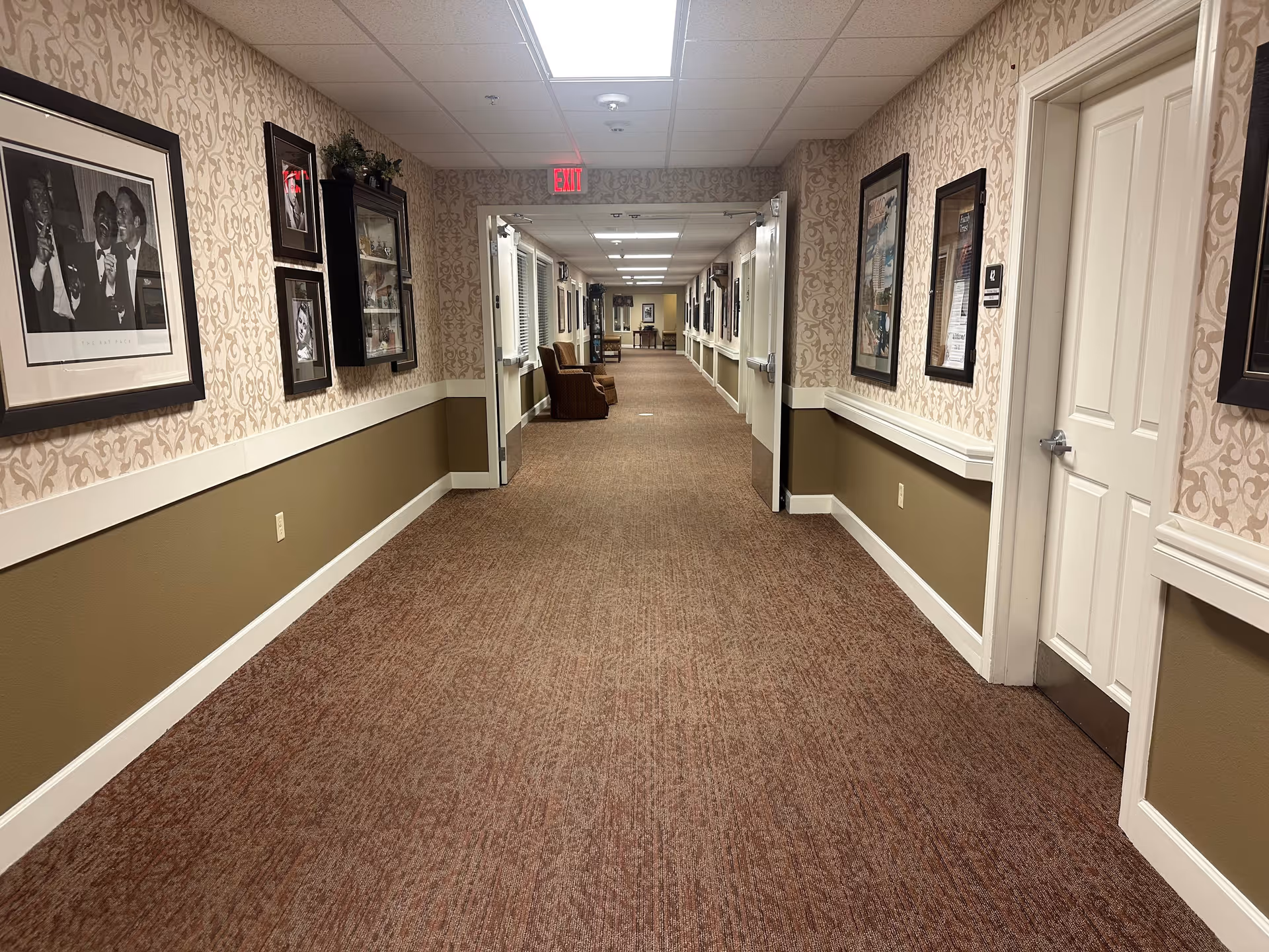 A long, carpeted hallway in a senior living facility with patterned wallpaper and framed pictures on the walls. There are handrails along both sides of the hallway and a few chairs placed near the far end. The ceiling has recessed lighting and an exit sign is visible above double doors at the end of the hallway.