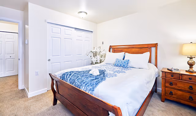 A bright bedroom with a wooden bed frame featuring white and blue bedding, a wooden nightstand with a lamp, and a closet with white sliding doors. The room has beige carpet and light-colored walls.