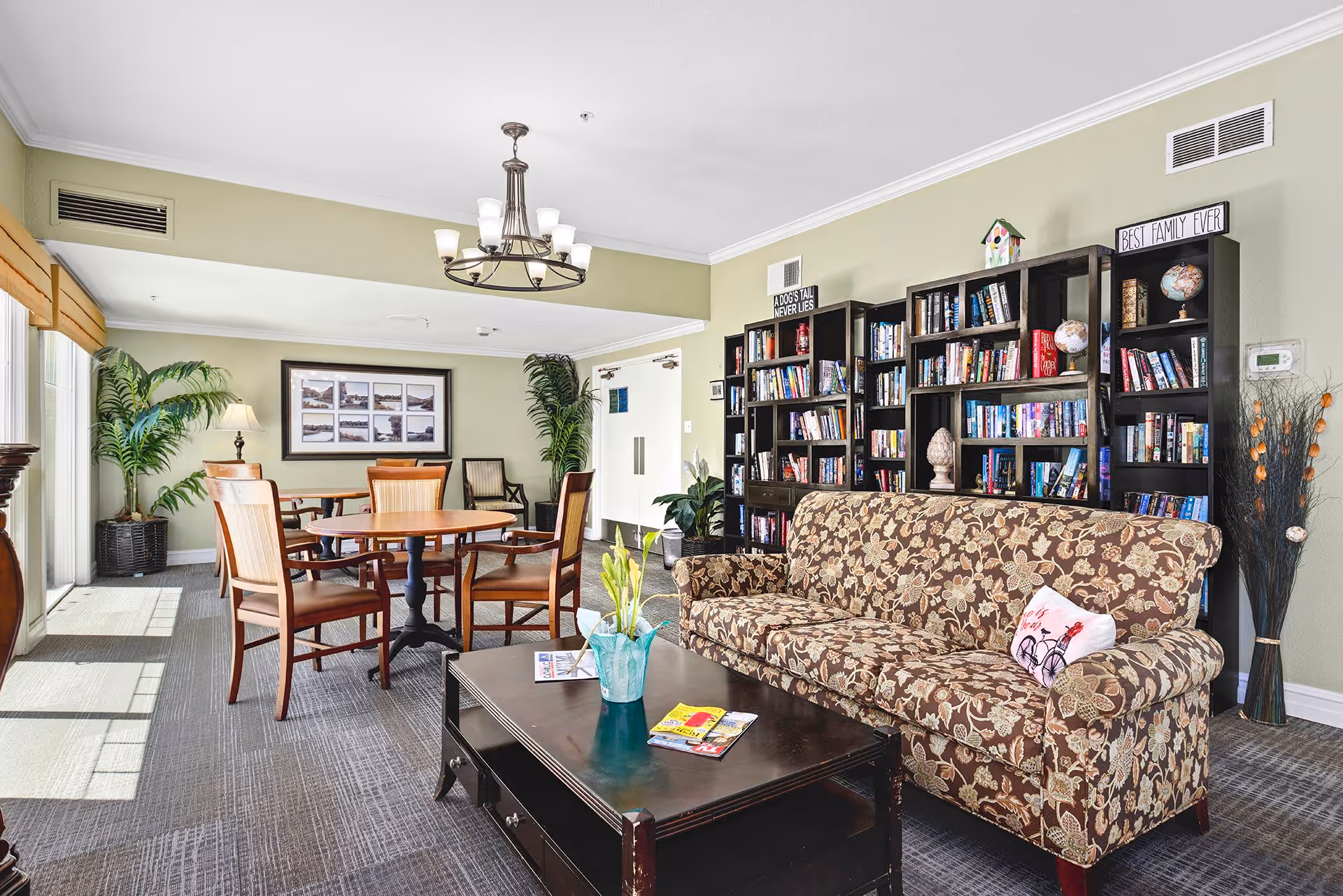 A bright and cozy living room area in Montecito Assisted Living featuring a floral patterned sofa with a decorative pillow, a dark wooden coffee table with magazines and a vase with flowers, a round wooden table with four chairs, large bookshelves filled with books and decorative items, potted plants, and a chandelier hanging from the ceiling.