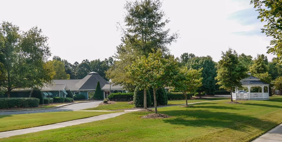 A well-maintained outdoor area of a senior living facility featuring a green lawn, several trees, a paved walkway, a white gazebo, and a building with a sloped roof in the background under a partly cloudy sky.