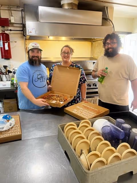 Three people standing in a kitchen. One person on the left is wearing a blue shirt and a cap, holding an open pizza box with a pizza inside. The person in the middle is wearing glasses and a patterned shirt, smiling. The person on the right is holding a green bottle and wearing a beige shirt. In the foreground, there is a metal tray with stacked plates and cups. The kitchen has a stove and a large exhaust hood in the background.